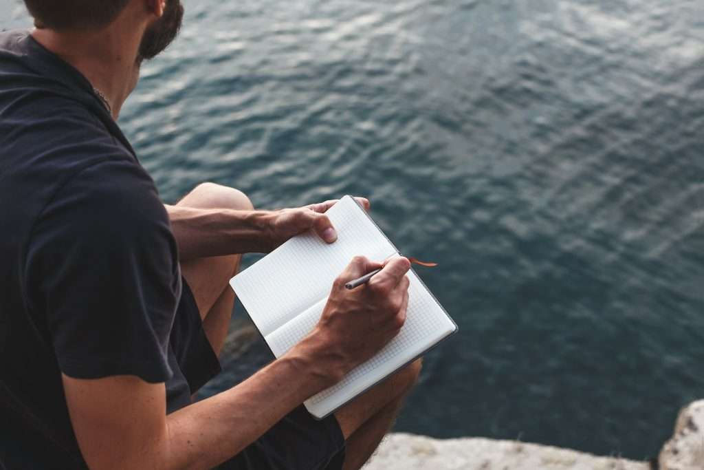 Brandheart Insights. Man writing in a notebook sitting on a rock over water.
